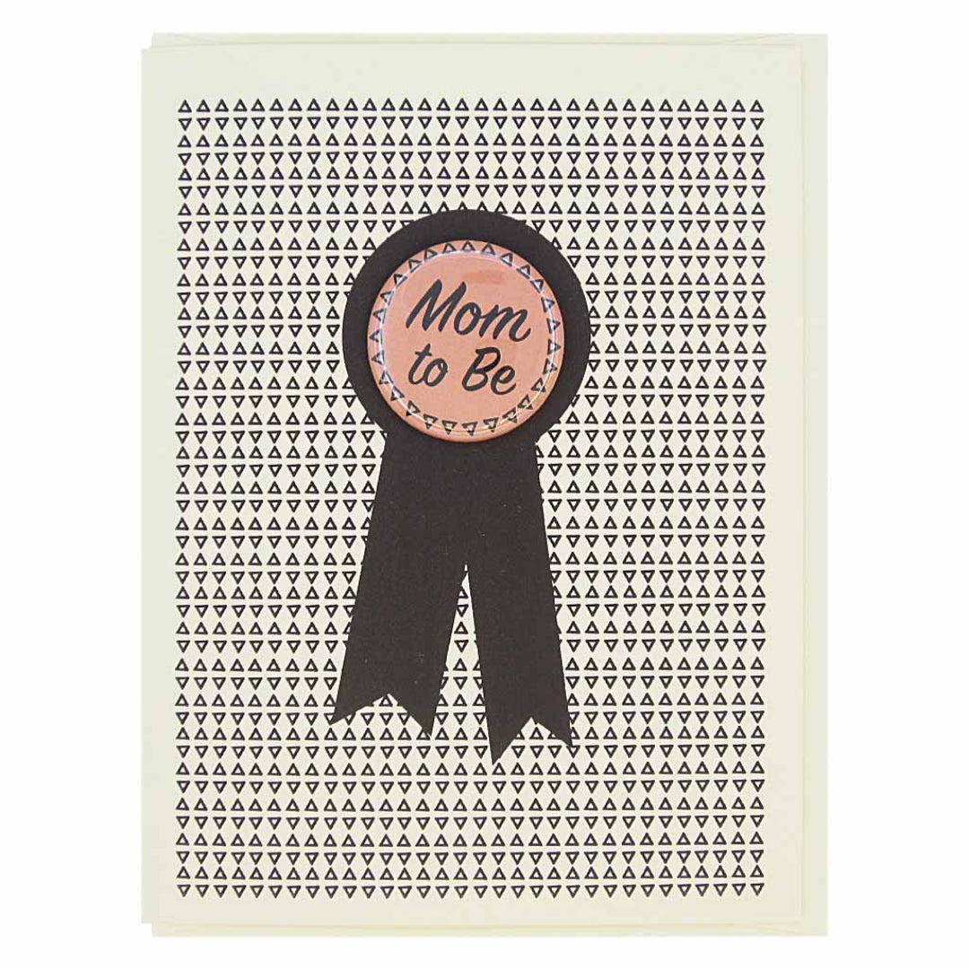 A congratulations card for a mom-to-be. The card has a white background with a pattern of small black triangles. In the center, there's a black ribbon shape with a round, light pink button on top that reads "Mom to Be" in an elegant script.