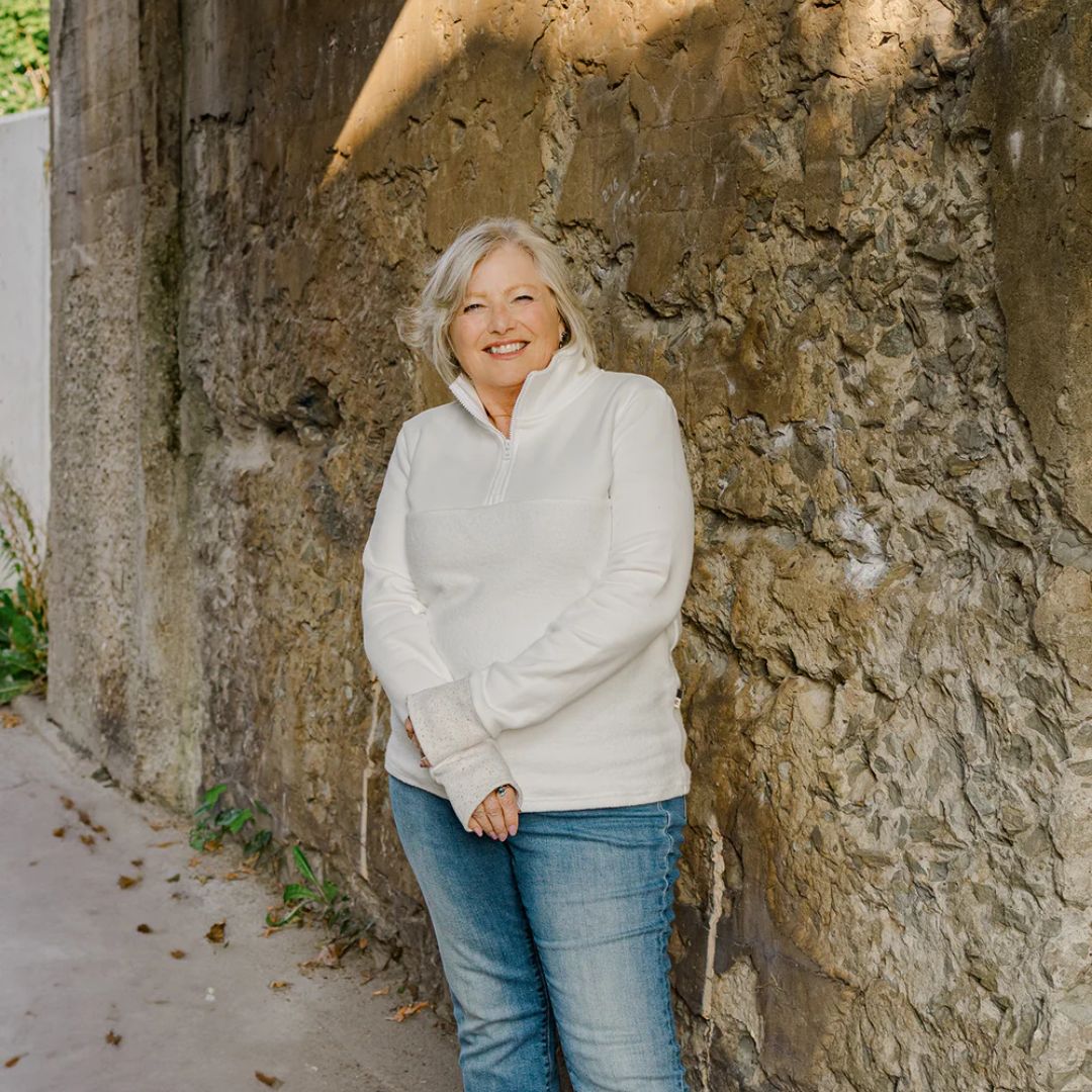 Woman in a white hoodie and blue jeans standing against a stone wall.