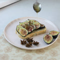 Person drizzling honey over figs on toast with a spoon on a white plate.