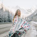 The Nibi Reversible Blanket by Mini Tipi, featuring bold Indigenous-inspired geometric patterns in teal, red, brown, and white, wrapped around a person in the mountains. This reversible and stylish blanket is available in Ottawa, Ontario, Canada.