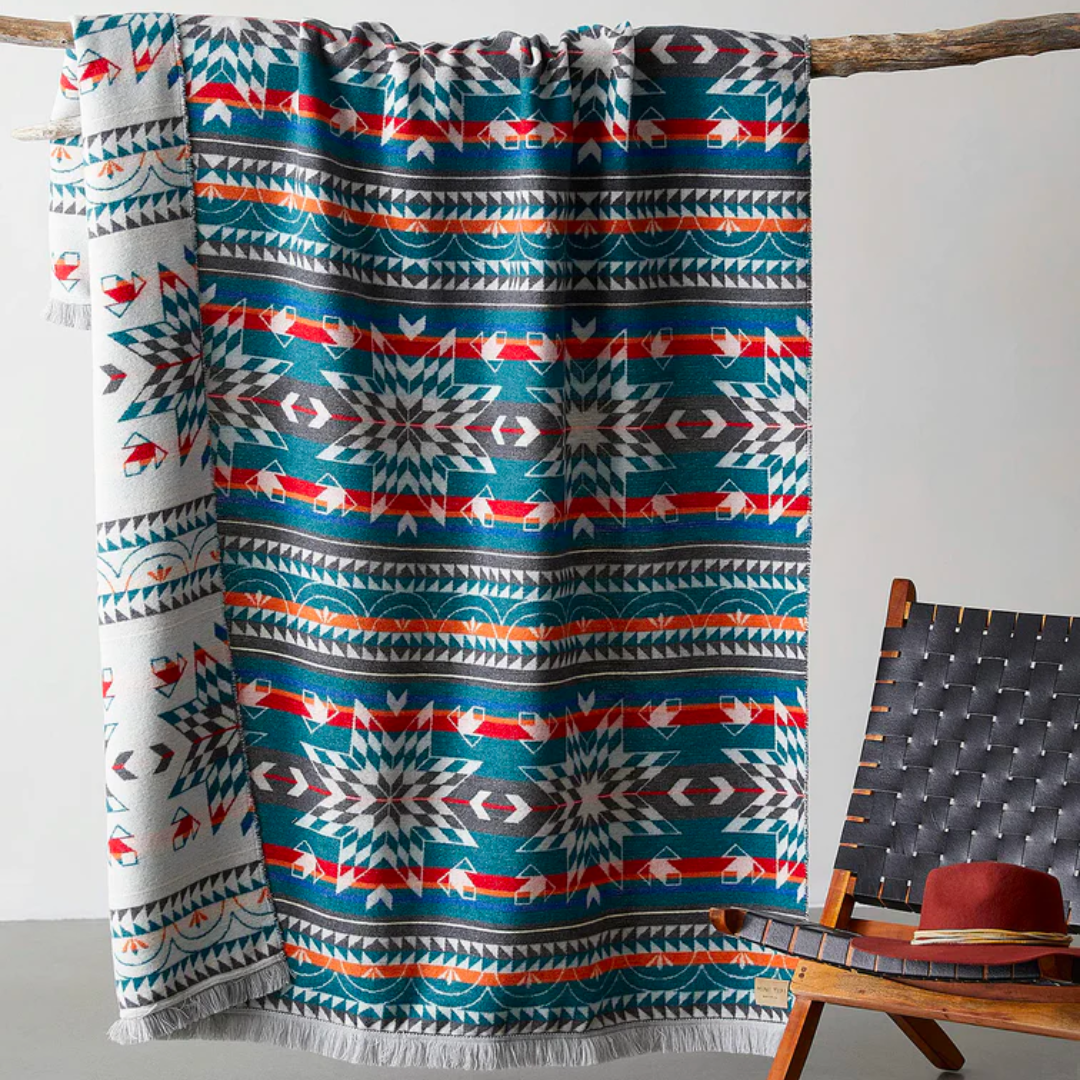 The Nibi Reversible Blanket by Mini Tipi, featuring bold Indigenous-inspired geometric patterns in teal, red, brown, and white, hanging from a natural branch. This reversible and stylish blanket is available in Ottawa, Ontario, Canada.