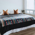 The Nibi Reversible Blanket by Mini Tipi, featuring bold Indigenous-inspired geometric patterns in teal, red, brown, and white, draped on a bed. This reversible and stylish blanket is available in Ottawa, Ontario, Canada.