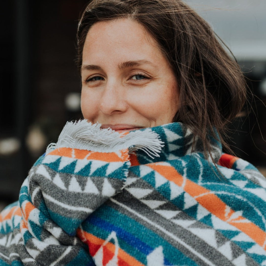 The Nibi Reversible Blanket by Mini Tipi, featuring bold Indigenous-inspired geometric patterns in teal, red, brown, and white, wrapped around one of the owners of MINITIPI. This reversible and stylish blanket is available in Ottawa, Ontario, Canada.