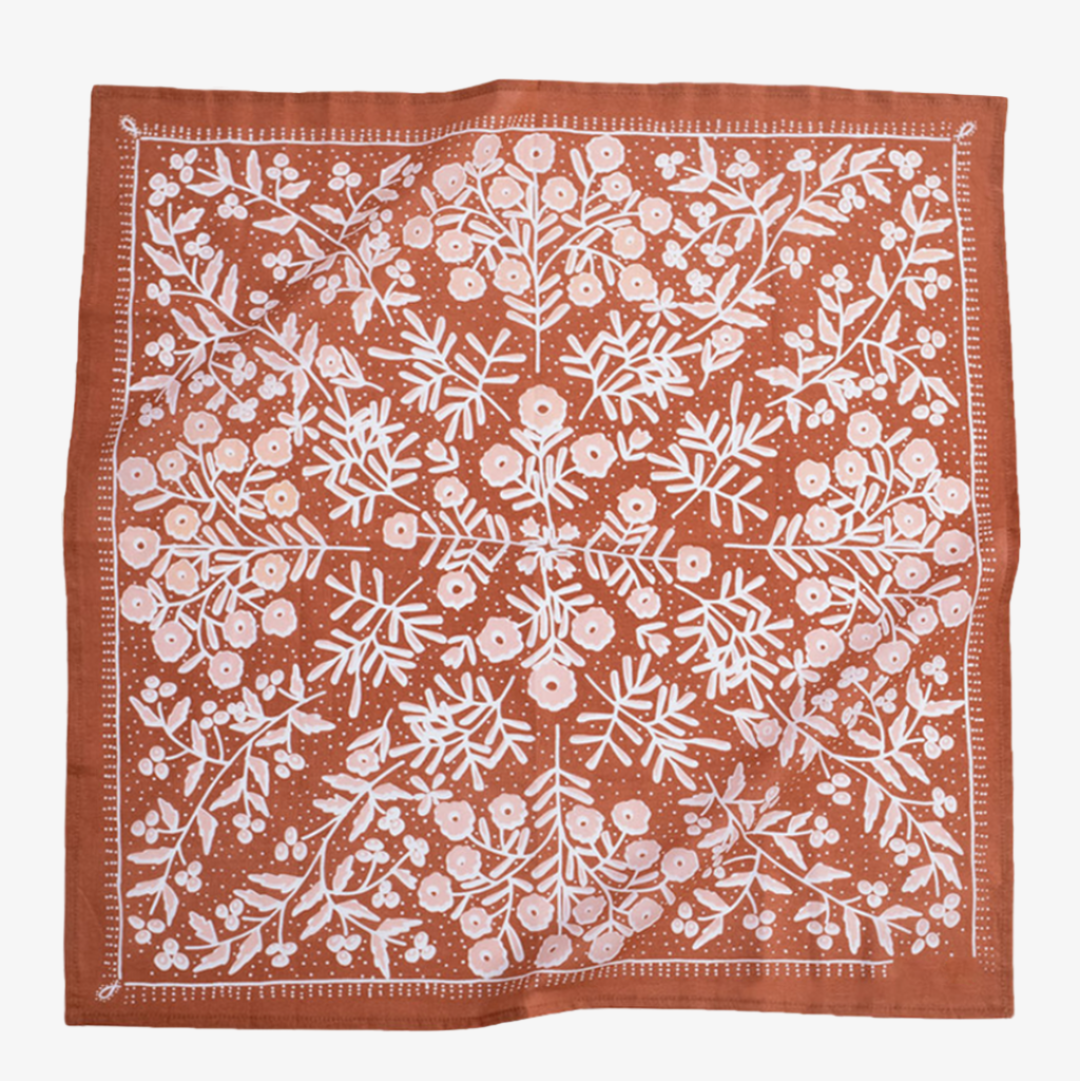 A terracotta-colored bandana with a floral pattern. The pattern features white flowers with pink centers and green leaves. The border of the bandana has a decorative, repeating pattern.