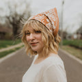 A terracotta-colored bandana with a floral pattern. The pattern features white flowers with pink centers and green leaves. The border of the bandana has a decorative, repeating pattern.
