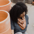 A terracotta-colored bandana with a floral pattern. The pattern features white flowers with pink centers and green leaves. The border of the bandana has a decorative, repeating pattern.