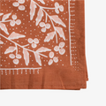 A terracotta-colored bandana with a floral pattern. The pattern features white flowers with pink centers and green leaves. The border of the bandana has a decorative, repeating pattern.