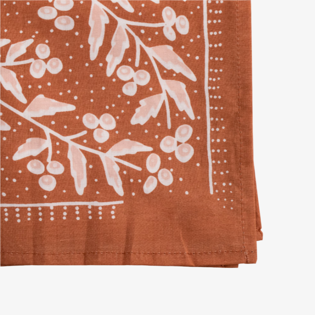 A terracotta-colored bandana with a floral pattern. The pattern features white flowers with pink centers and green leaves. The border of the bandana has a decorative, repeating pattern.