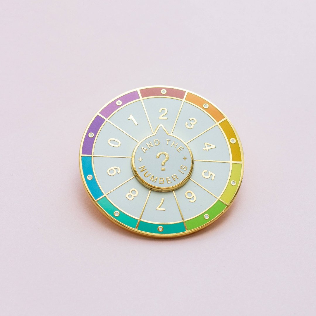 A circular pin with a colorful wheel featuring numbers from 0 to 9. The phrase "And the Numbers" is written in the center.
