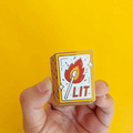 A hand holding a gold-colored enamel pin shaped like a matchbox. The matchbox has a lit match with a flame and the word "LIT" written on it.