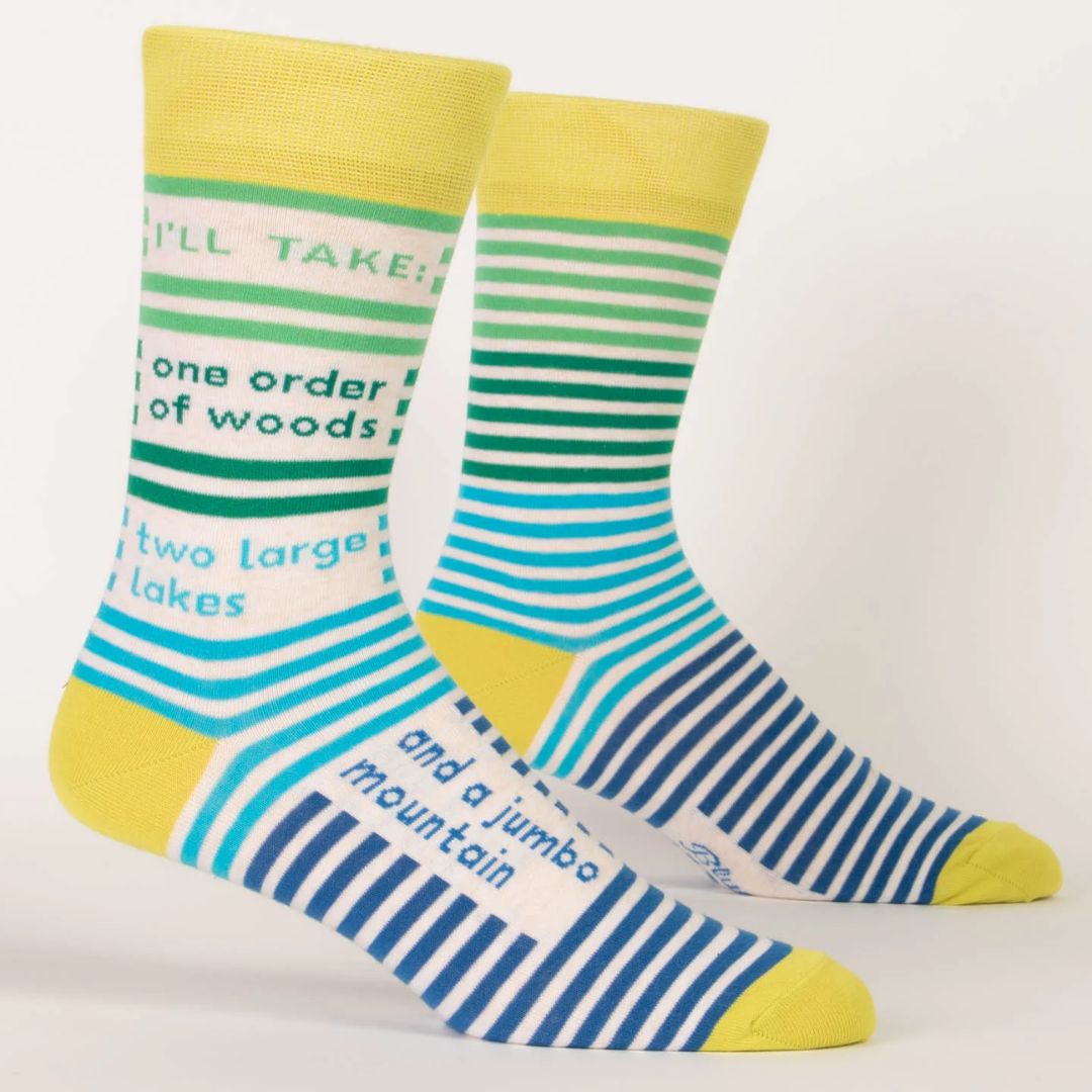 pair of men's crew socks with a vibrant stripe pattern. One sock features wide horizontal green and blue stripes, while the other has blue and white stripes on the foot and green and white stripes on the calf. Text on the socks reads "I'LL TAKE: one order of woods two large lakes and a jumbo mountain."