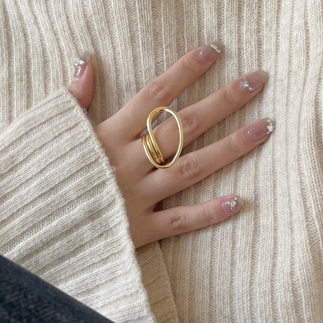 A gold-plated geometric open statement twist ring, featuring a unique, flowing design, worn on a person's finger against a light-colored sweater. This is a modern and bold ring, available in Ottawa, Ontario, Canada.