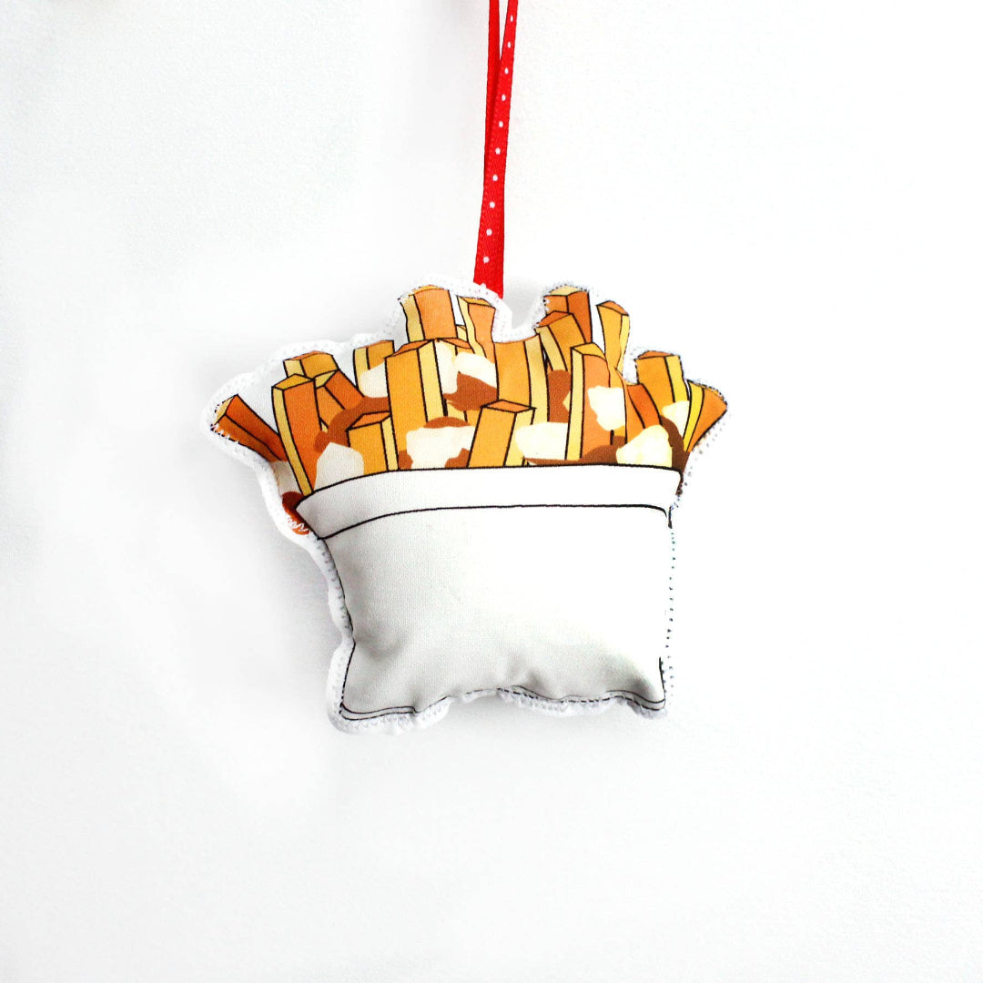 A close-up, high-angle flat lay shot on a plain white background features a soft, plush Christmas ornament shaped like a serving of poutine. The illustration shows a white cup filled with golden-brown french fries and a small amount of gravy or cheese curds. A red polka-dot ribbon loop is attached to the top for hanging.