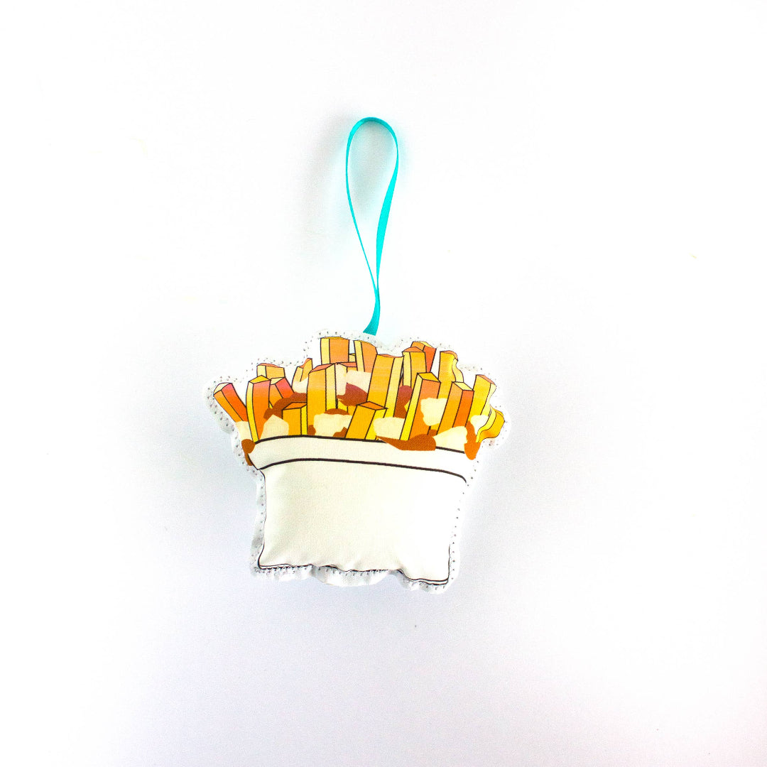 A close-up, high-angle flat lay shot on a plain white background features a soft, plush Christmas ornament shaped like a serving of poutine. The illustration shows a white cup filled with golden-brown french fries and a small amount of gravy or cheese curds. A red polka-dot ribbon loop is attached to the top for hanging.