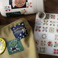 Decorative items including a mug with stickers, a green patch, and a card with floral designs on a wooden surface.