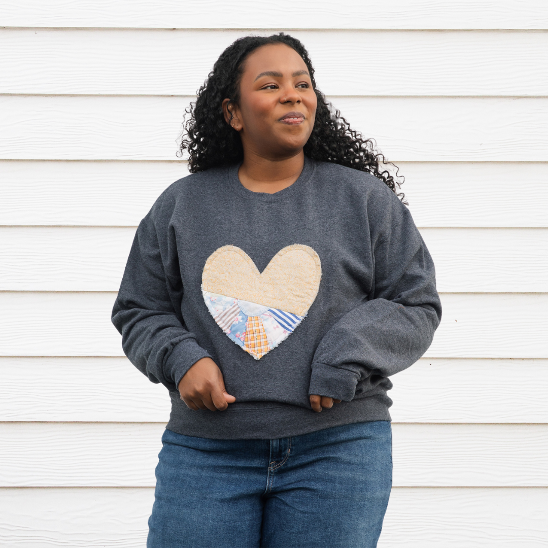 a grey sweatshirt with a large, heart-shaped patch. The patch is made from various fabrics with various patterns, sewn together in a patchwork style.