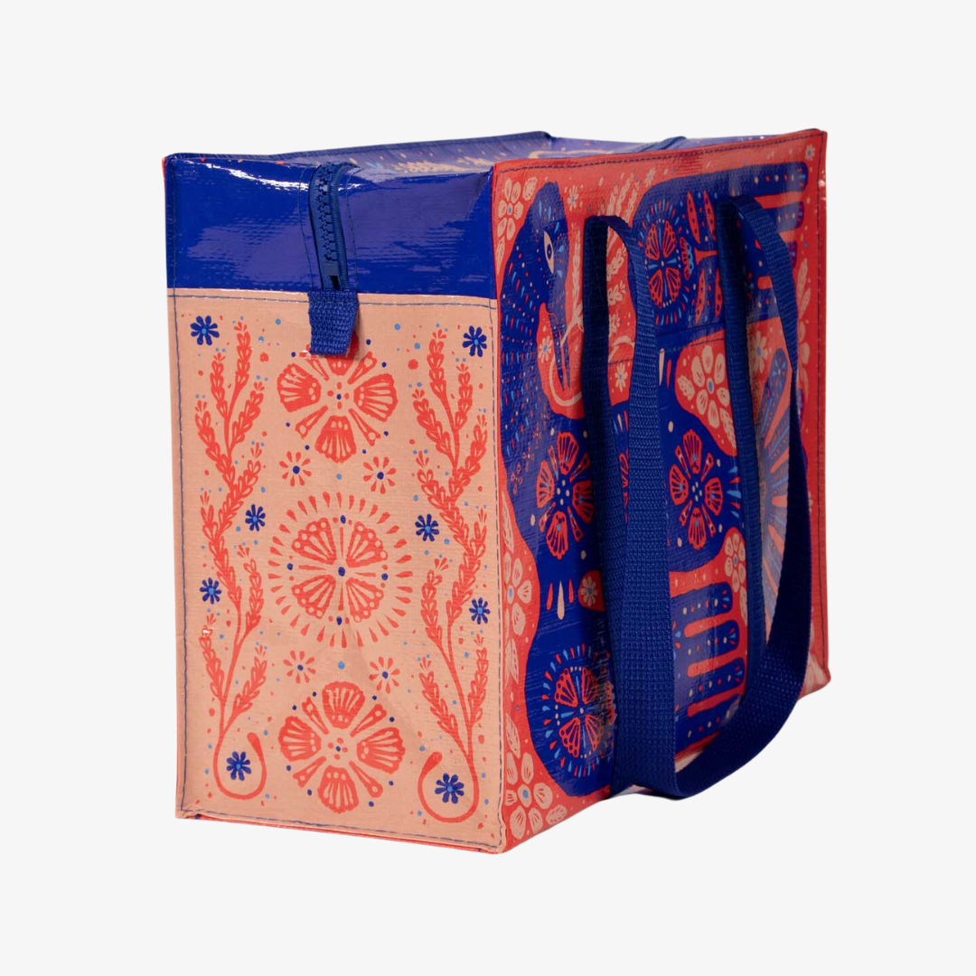 A reusable shoulder tote bag with a vibrant and colorful design. The bag is made from recycled materials and features a large, folk-art style bird surrounded by flowers and plants. The bird is primarily blue with red accents, while the flowers are in shades of pink, orange, and yellow. The bag has sturdy blue handles.