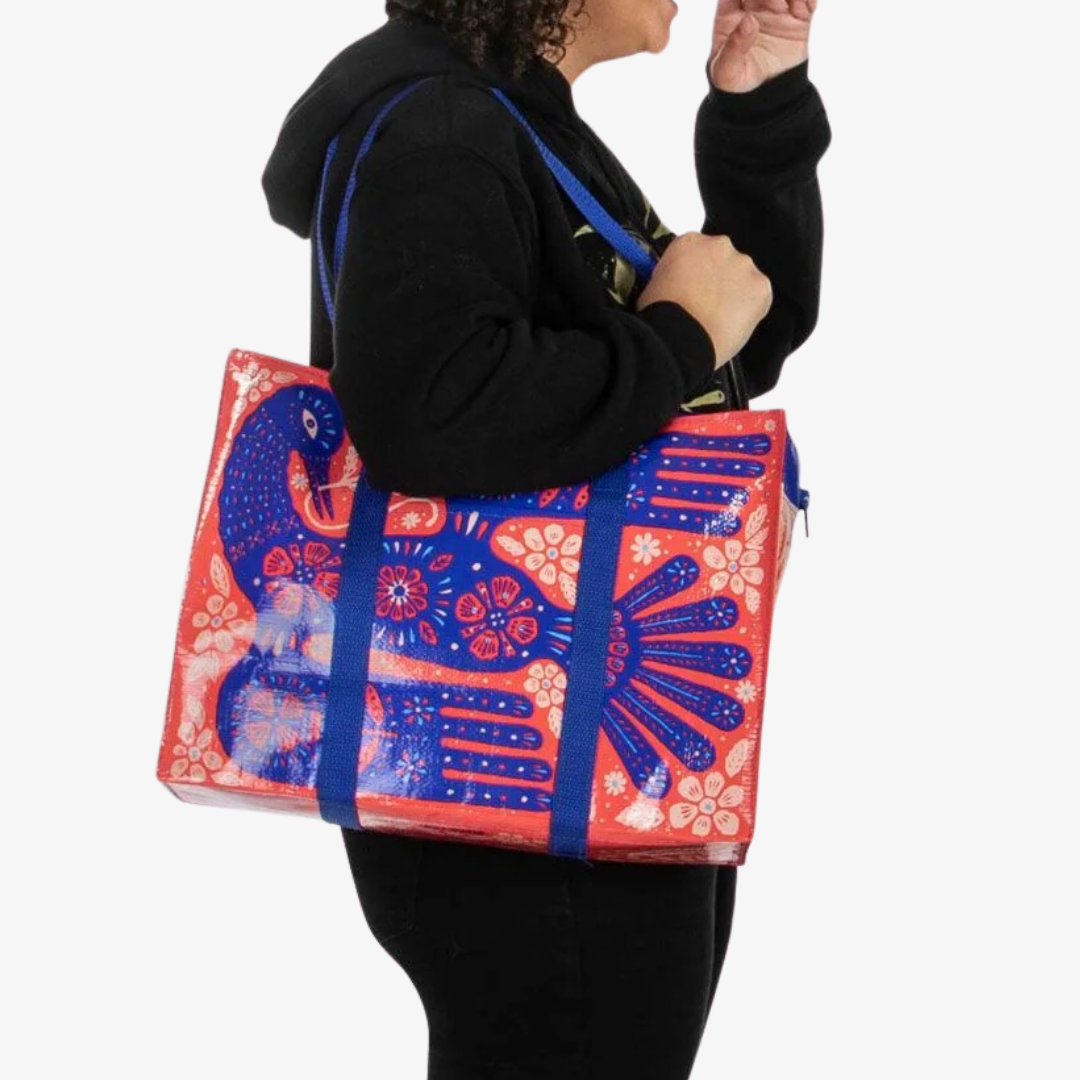 A reusable shoulder tote bag with a vibrant and colorful design. The bag is made from recycled materials and features a large, folk-art style bird surrounded by flowers and plants. The bird is primarily blue with red accents, while the flowers are in shades of pink, orange, and yellow. The bag has sturdy blue handles.