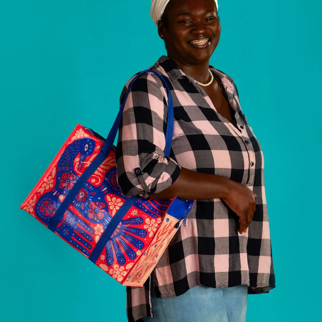 A reusable shoulder tote bag with a vibrant and colorful design. The bag is made from recycled materials and features a large, folk-art style bird surrounded by flowers and plants. The bird is primarily blue with red accents, while the flowers are in shades of pink, orange, and yellow. The bag has sturdy blue handles.