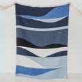 The River Reversible Blanket by Mini Tipi, featuring flowing waves of blue, grey, black, and white, held up by two hands. This reversible blanket is available in Ottawa, Ontario, Canada.