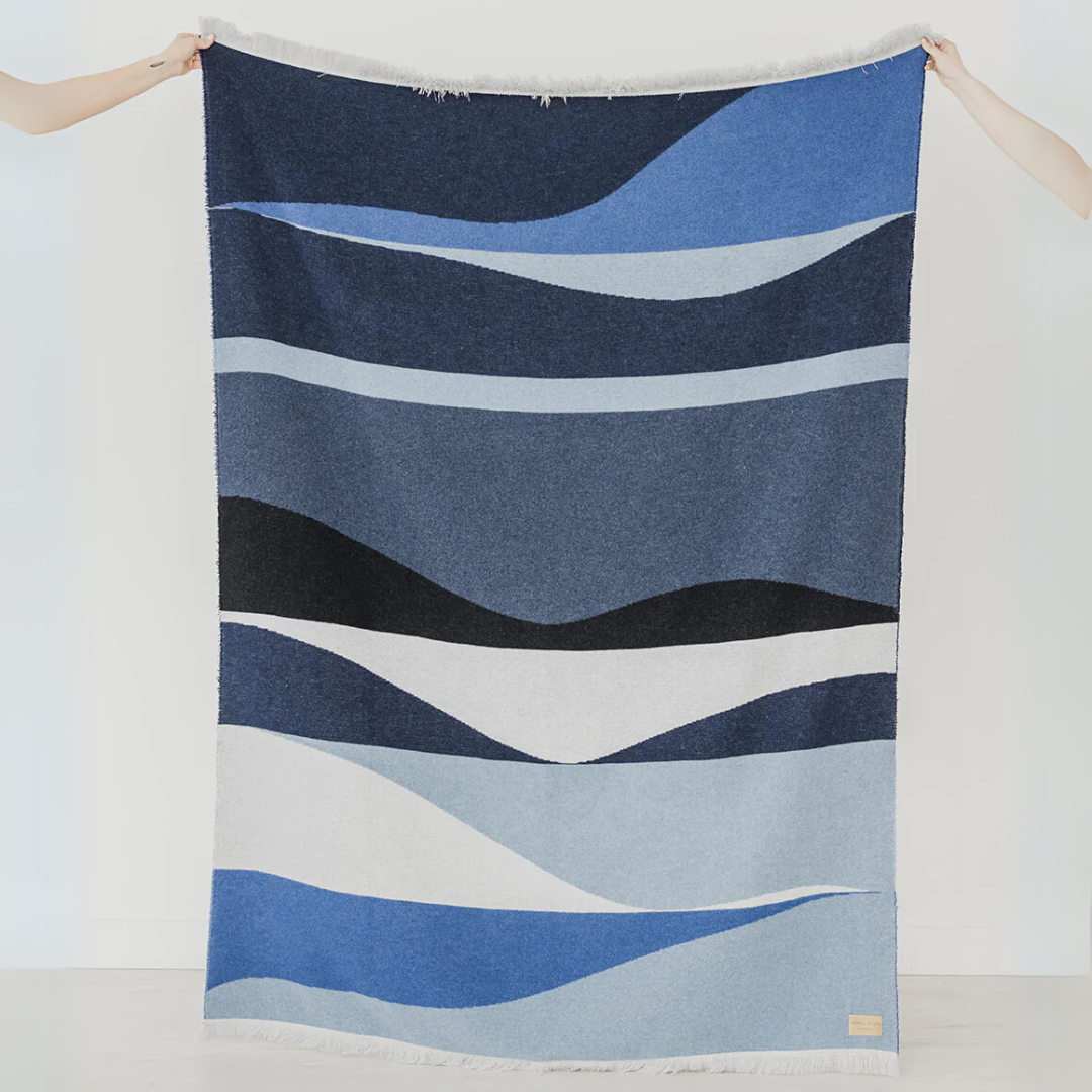 The River Reversible Blanket by Mini Tipi, featuring flowing waves of blue, grey, black, and white, held up by two hands. This reversible blanket is available in Ottawa, Ontario, Canada.