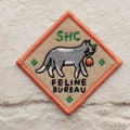 A diamond-shaped embroidered patch with an orange border. It features a gray cat carrying a red flower in its mouth, walking to the right. The text "SHC" is in the top left corner and "FELINE BUREAU" is below the cat. The patch has four small stars at the corners.
