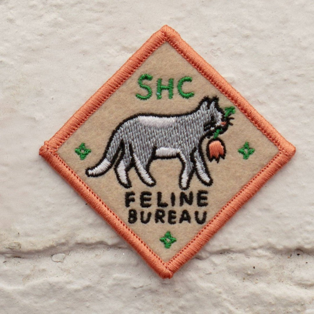 A diamond-shaped embroidered patch with an orange border. It features a gray cat carrying a red flower in its mouth, walking to the right. The text "SHC" is in the top left corner and "FELINE BUREAU" is below the cat. The patch has four small stars at the corners.