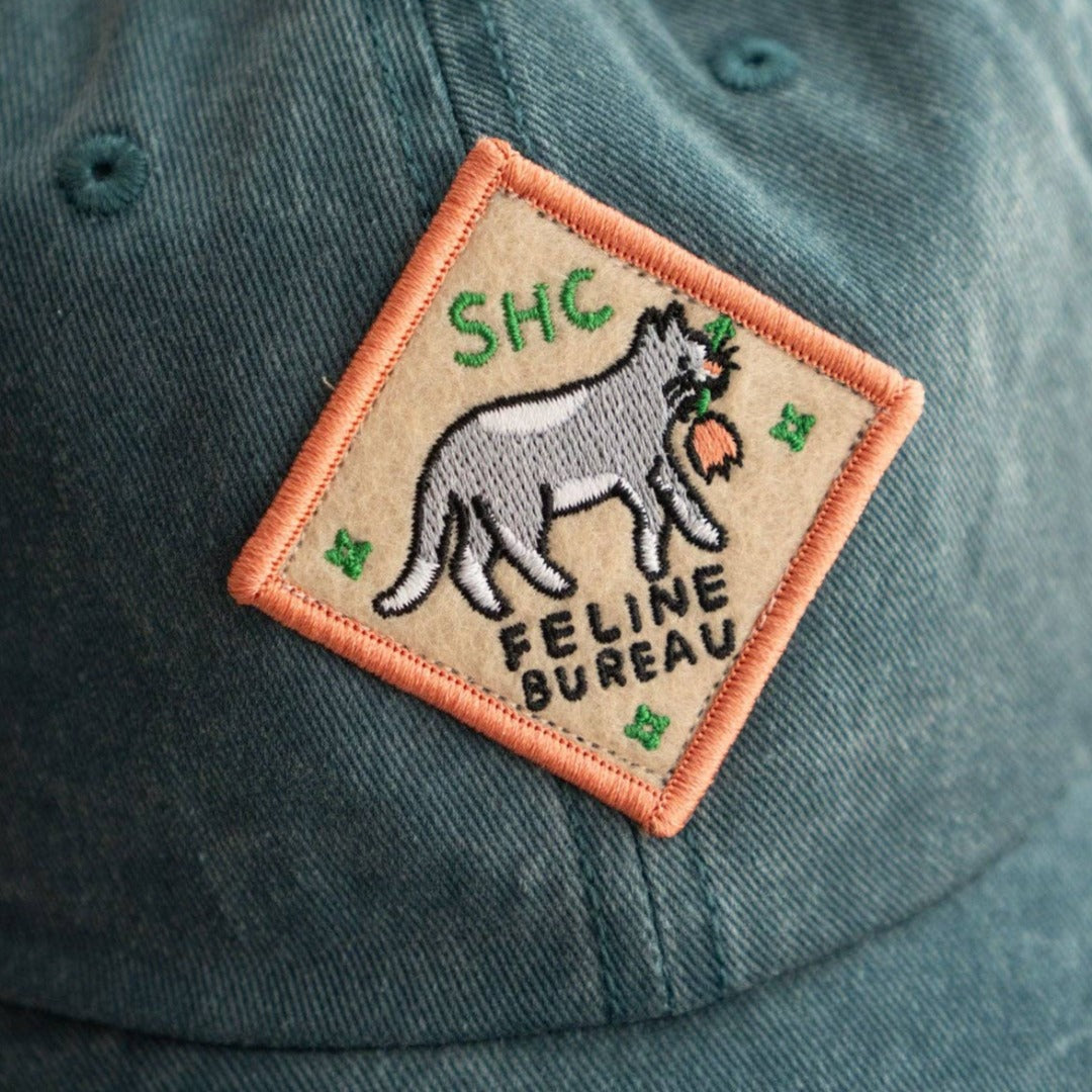 A diamond-shaped embroidered patch with an orange border. It features a gray cat carrying a red flower in its mouth, walking to the right. The text "SHC" is in the top left corner and "FELINE BUREAU" is below the cat. The patch has four small stars at the corners.