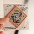 A diamond-shaped embroidered patch with an orange border. It features a gray cat carrying a red flower in its mouth, walking to the right. The text "SHC" is in the top left corner and "FELINE BUREAU" is below the cat. The patch has four small stars at the corners.