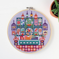 An overhead shot of a finished cross-stitch piece in a wooden embroidery hoop, displayed on a white surface with a potted succulent in the top right corner. The cross-stitch design features a colourful and intricate illustration of a shop interior with shelves, books, plants, and decorative items.