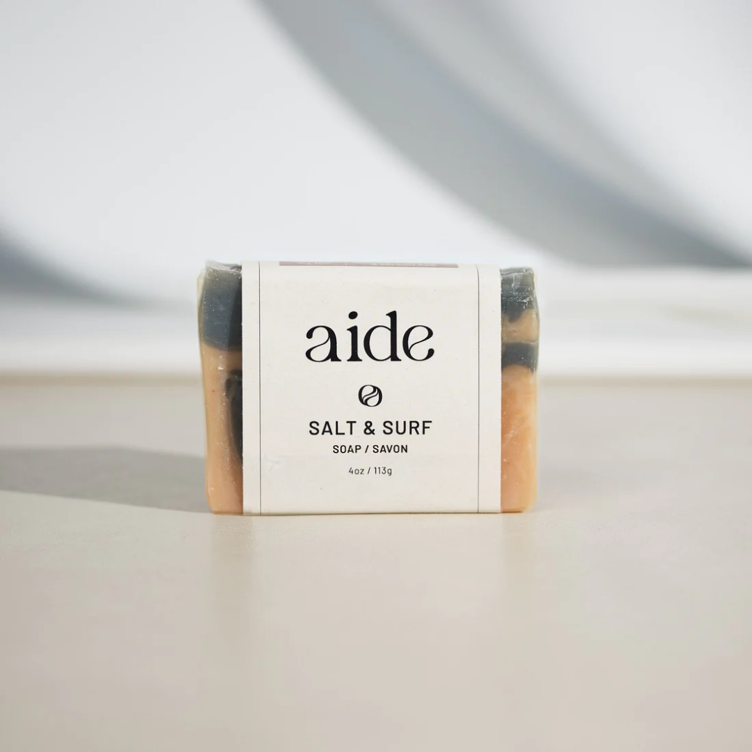 A bar of Aide Bodycare Salt & Surf Soap, featuring a light orange and dark gray swirled pattern and a paper label with the 'aide' logo and 'Salt & Surf' text, displayed on a light-colored surface with a shadow overlay. The soap is natural and handmade, available in Ottawa, Ontario, Canada.
