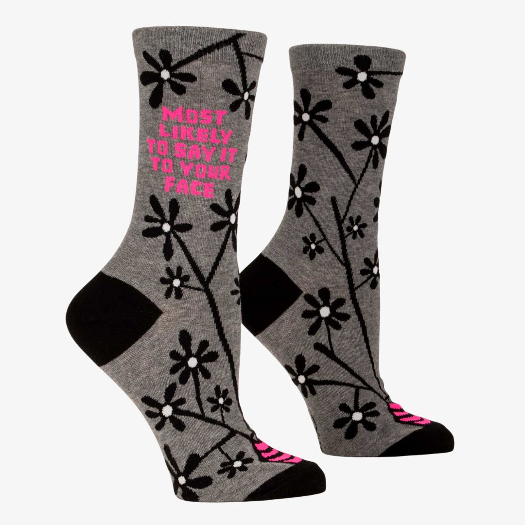A pair of women's crew socks with a grey background and a pattern of black and white flowers on branches. One sock has text in bright pink that reads "MOST LIKELY TO SAY IT TO YOUR FACE".