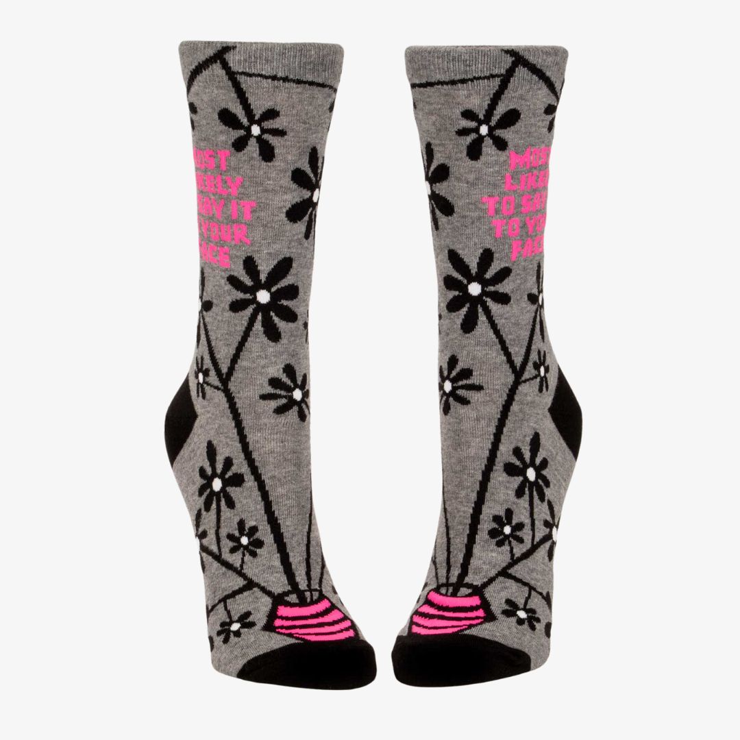 A pair of women's crew socks with a grey background and a pattern of black and white flowers on branches. One sock has text in bright pink that reads "MOST LIKELY TO SAY IT TO YOUR FACE".