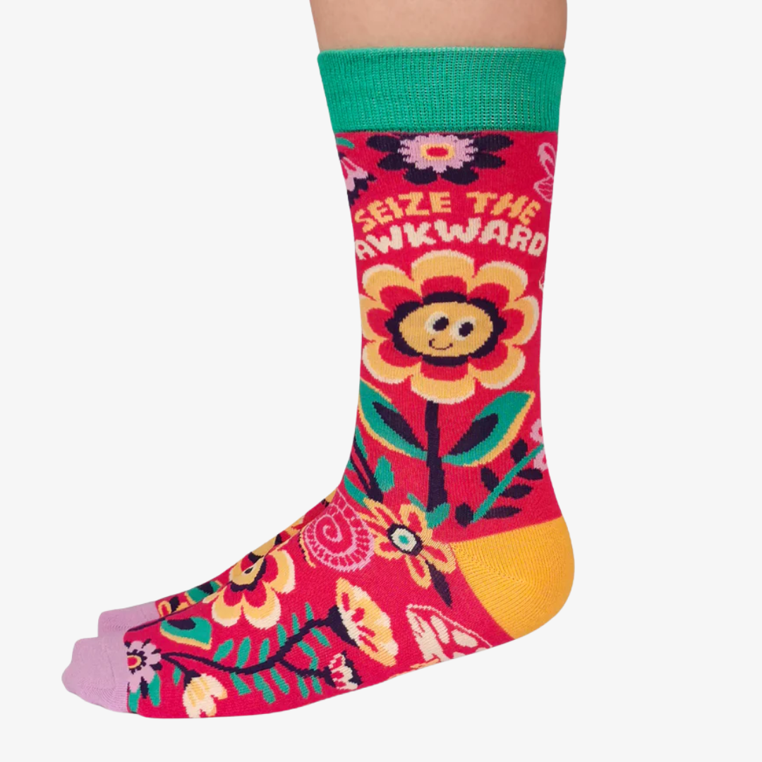 A pair of colourful women's crew socks with a red background featuring a whimsical floral pattern in shades of yellow, pink, green, and white. One sock displays the text "SEIZE THE AWKWARD" in white letters around a smiling yellow flower. The toe of the sock is light purple, the heel is yellow, and the cuff is bright green.