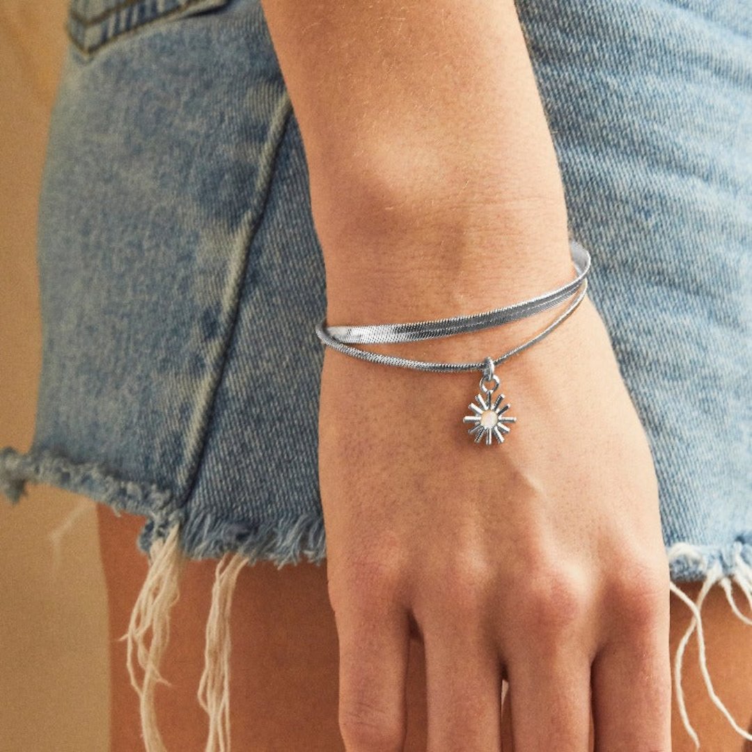 A set of two silver bracelets, one layered over the other, with a sun charm hanging from the bottom. The bracelets are displayed on a pink card with the brand name "Foxy Originals."