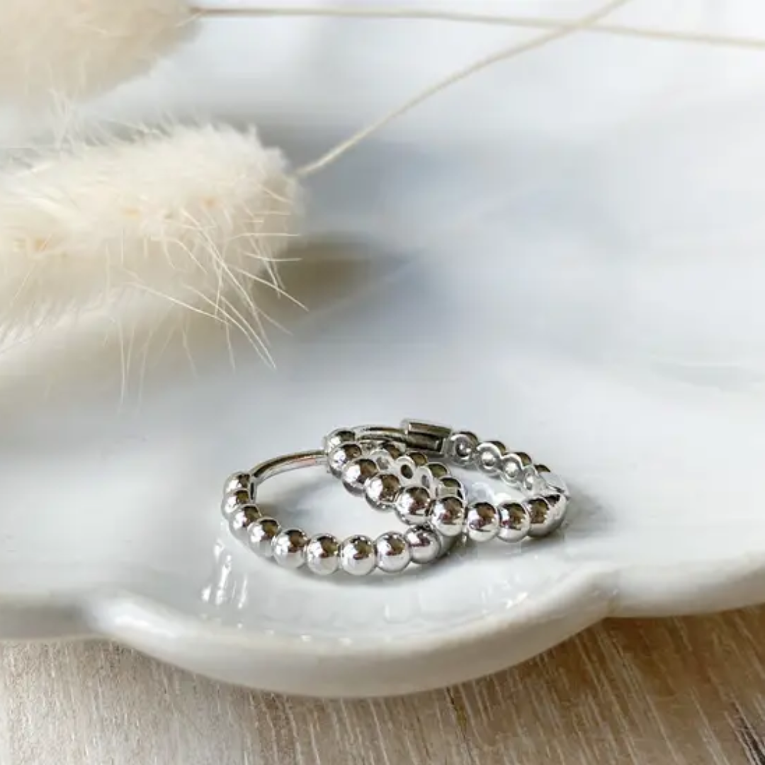A pair of 925 sterling silver ball hoop earrings, featuring a beaded design, displayed on a white textured surface with white floral accents. The earrings are dainty and high-quality, available in Ottawa, Ontario, Canada.
