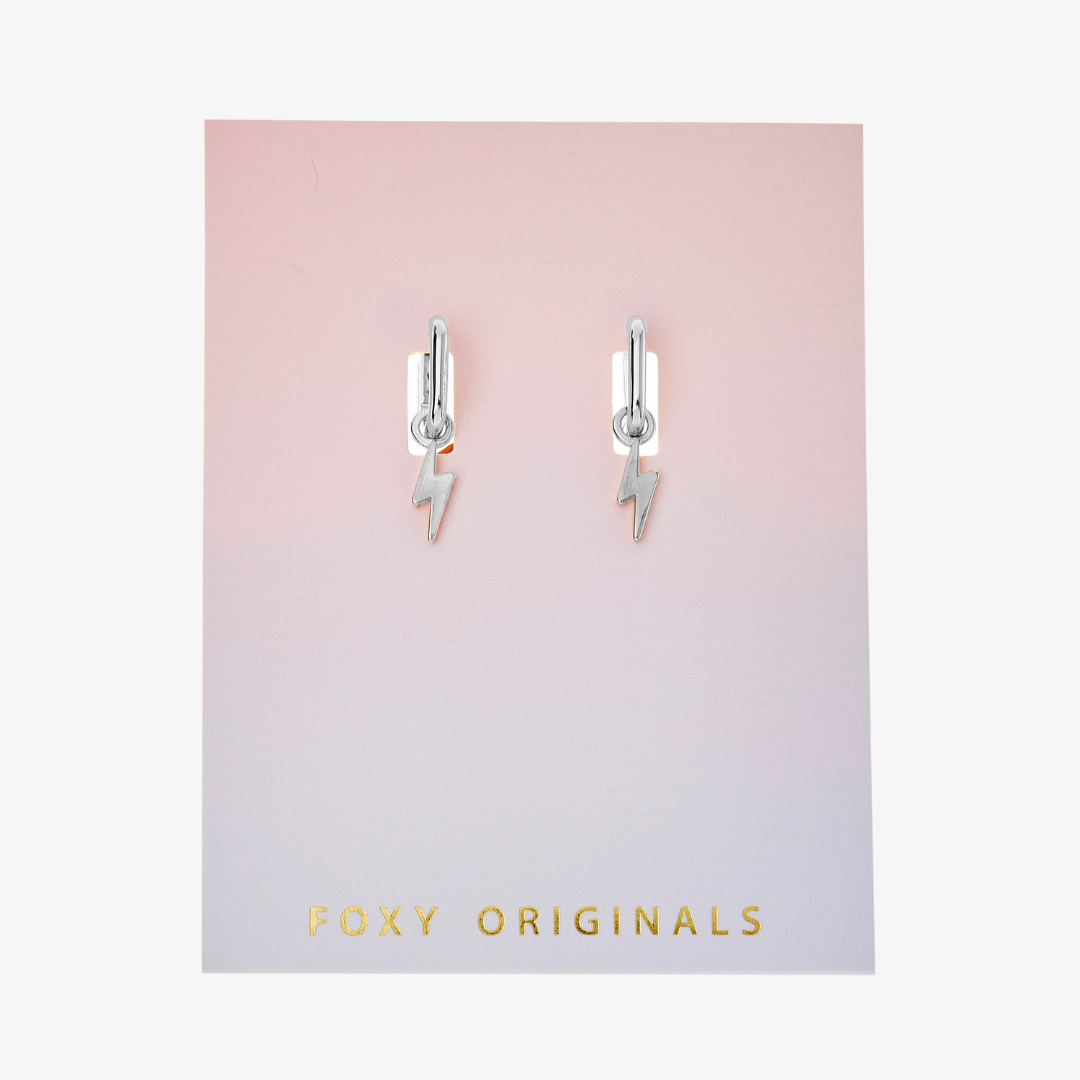 A pair of silver earrings with lightning bolt charms on a pink background. The earrings are displayed on a card with the brand name "Foxy Originals."