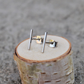A pair of silver bar stud earrings resting on a small wooden disc. The earrings are simple and minimalist, with a single straight bar for each earring.