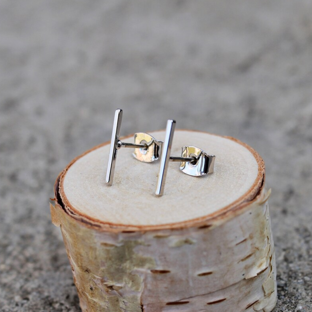 A pair of silver bar stud earrings resting on a small wooden disc. The earrings are simple and minimalist, with a single straight bar for each earring.