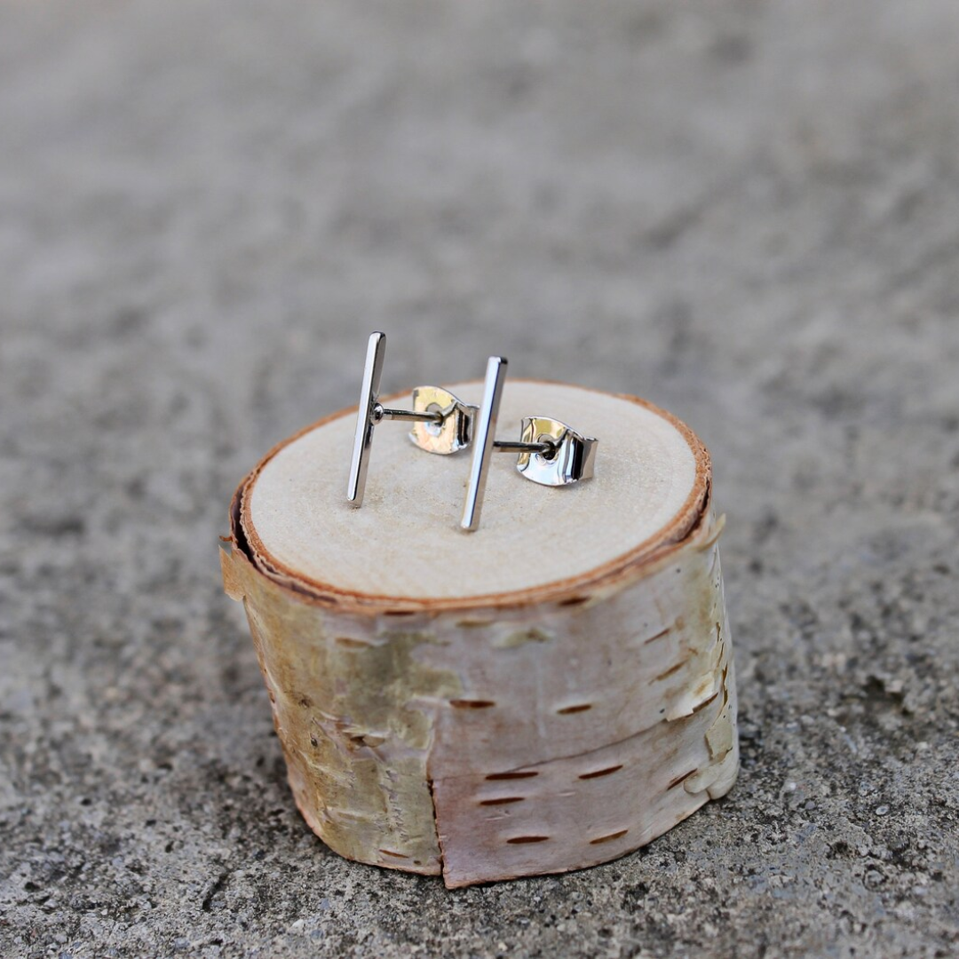 A pair of silver bar stud earrings resting on a small wooden disc. The earrings are simple and minimalist, with a single straight bar for each earring.