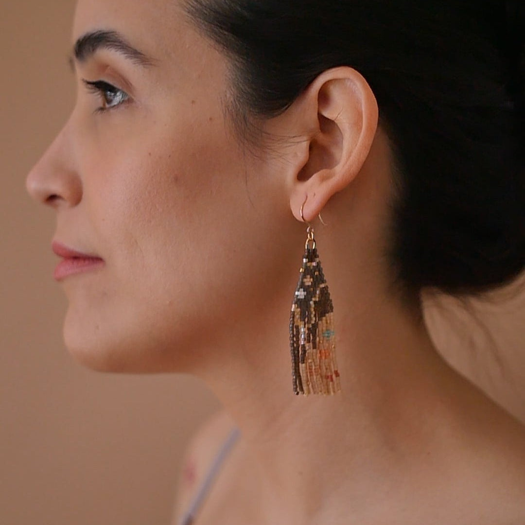 A pair of long, triangular tapestry-style earrings hand-stitched with small Japanese glass beads to depict a stylized face with light peach skin, blue eyes, coral cheeks, and a red mouth, framed by dark brown and grey hair with gold bead accents. They hang from delicate 14 KT gold filled fishhook ear wires.
