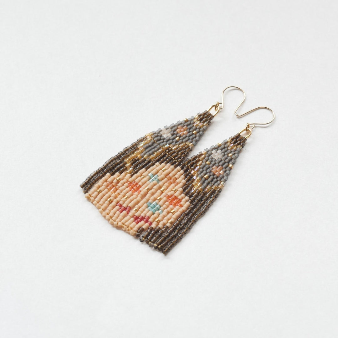A pair of long, triangular tapestry-style earrings hand-stitched with small Japanese glass beads to depict a stylized face with light peach skin, blue eyes, coral cheeks, and a red mouth, framed by dark brown and grey hair with gold bead accents. They hang from delicate 14 KT gold filled fishhook ear wires.