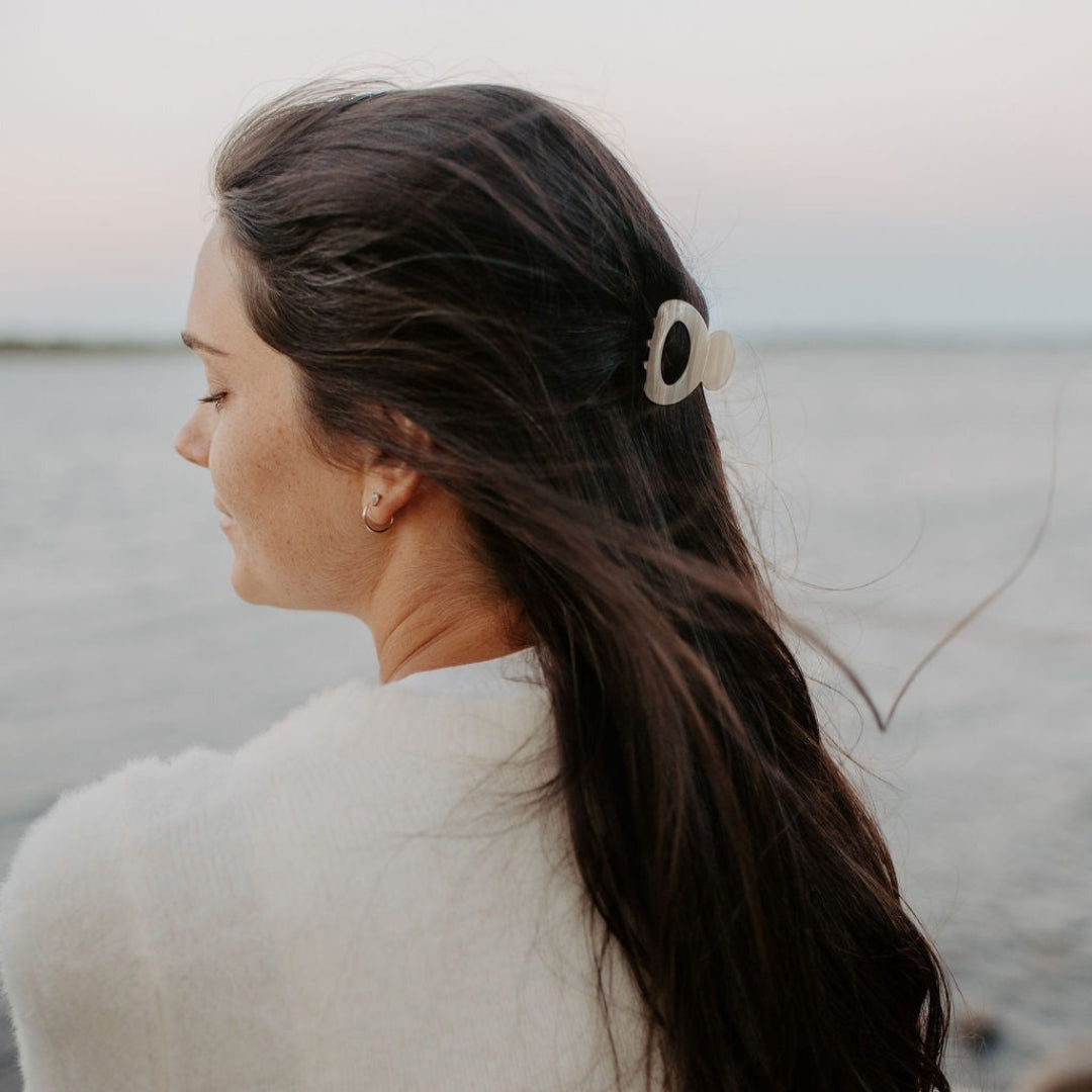 Woman with long dark hair wearing a white fluffy jacket by a body of water.