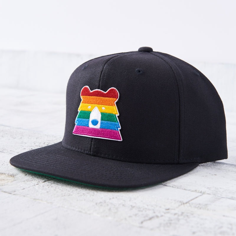 A black snapback cap with a flat green brim. The front features a unique bear face patch in the colours of the rainbow pride flag. Shop this at Cheerfully Made, Canadian-made gift shop.
