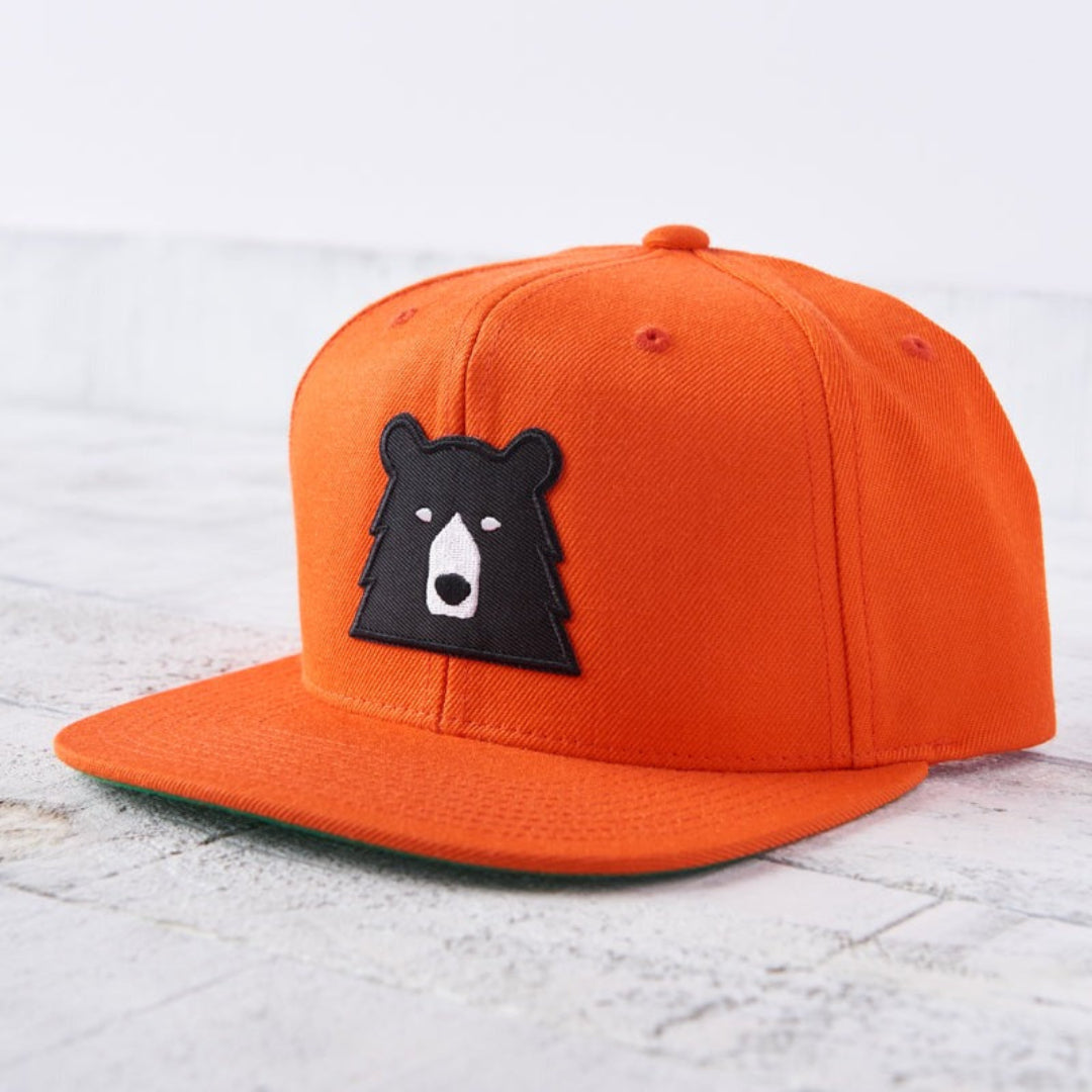 A bright orange (blaze) snapback cap with a flat green brim. The front features a black bear face patch with white eyes and snout. Shop this at Cheerfully Made, Canadian-made gift shop.