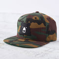 A camouflage snapback cap with a flat green brim. The front features a black bear face patch with white eyes and snout. Shop this at Cheerfully Made, Canadian-made gift shop.