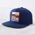 A navy blue snapback cap with a flat green brim. The front features a square patch with an orange sun, orange and brown waves, and green trees with "NORTH STANDARD" text in white. Shop this at Cheerfully Made, Canadian-made gift shop.
