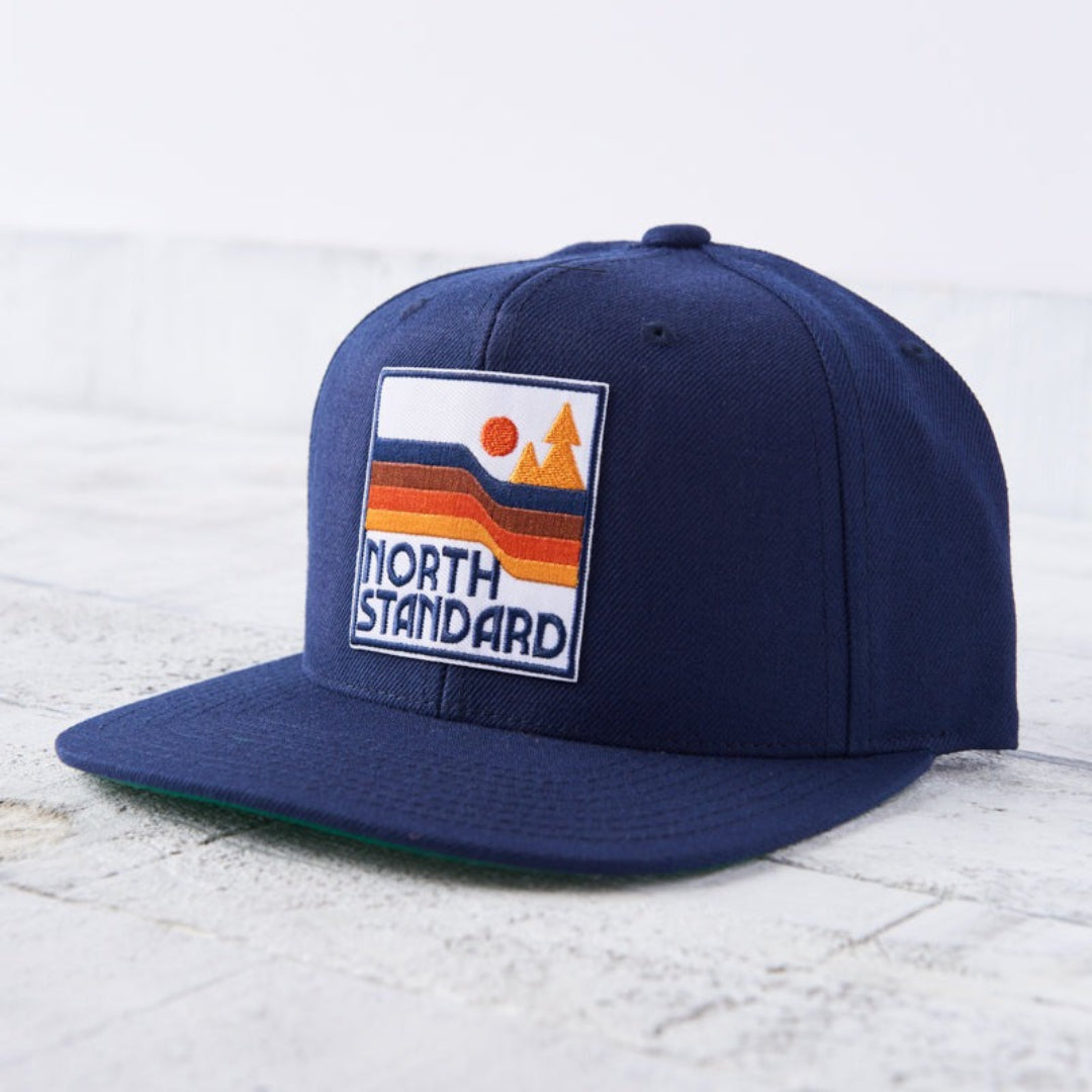 A navy blue snapback cap with a flat green brim. The front features a square patch with an orange sun, orange and brown waves, and green trees with "NORTH STANDARD" text in white. Shop this at Cheerfully Made, Canadian-made gift shop.