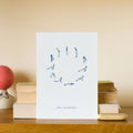 White card with blue illustrations of yoga poses on a wooden surface with books and a pink object.