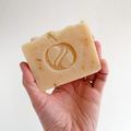A rectangular bar of soap with a light beige color. There is a circular logo embossed on the center of the soap.